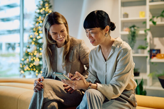 Diverse business women smiling, discussing project on smartphone, teamwork during holiday season in modern office setting - Powered by Adobe