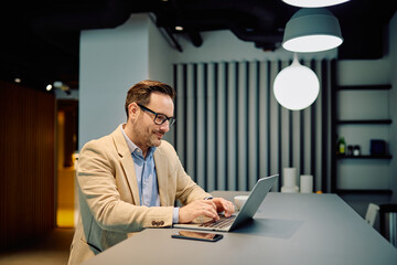 Smiling businessman in smart casual wear sitting at a desk, typing on laptop, working in a contemporary office