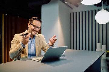 Smiling businessman working remotely, having a voice call on his smartphone while looking at a laptop in a modern office