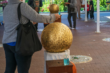 Visitor applies gold leaf onto a sacred golden sphere at a Thai Buddhist temple, surrounded by other worshippers.