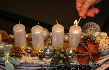 Hand lighting the first candle on an Advent wreath decorated with ornaments, pine cones and festive elements. Warm holiday atmosphere and Christmas tradition at home.