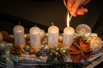 Hand lighting the first candle on an Advent wreath decorated with ornaments, pine cones and festive elements. Warm holiday atmosphere and Christmas tradition at home.