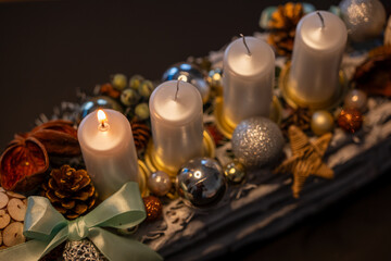 Advent wreath with four white candles, one lit for the first Advent Sunday. Festive Christmas decoration with ornaments, pine cones and natural elements in warm candlelight.