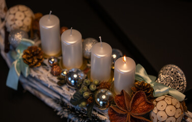 Advent wreath with four white candles, one lit for the first Advent Sunday. Festive Christmas decoration with ornaments, pine cones and natural elements in warm candlelight.