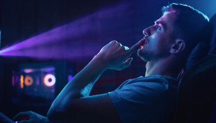 Young man focused and thinking while gaming or working on a computer in a dark room with purple light.