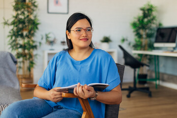 A thoughtful woman sits with her book in a comfortable armchair, set against a bright room backdrop, embodying coziness and creativity, enhanced by modern design elements.