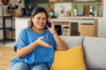 A happy woman engages in a phone call, smiling widely while seated on a couch with a yellow accent pillow, showcasing a moment of joy and connection in her stylish home.