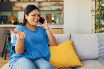A young woman smiling while talking on the phone, sitting comfortably on a light-colored couch, creating a warm and inviting atmosphere in her stylish home environment.