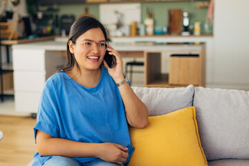 A young woman wearing glasses smiles while chatting on the phone in a modern, cozy living room setting filled with plants and warm colors that emphasize comfort and relaxation.