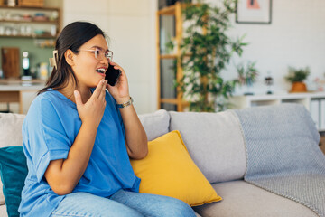 The young woman expresses joy and enthusiasm while on a phone call, showcasing a lively and cheerful atmosphere in her well-decorated living space with comfortable hues.