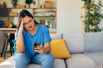A distressed woman is seen holding her phone in her comfortable home space, reflecting the emotional toll of modern communication and unexpected messages.