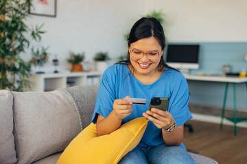 An excited woman browses her smartphone for online deals while holding a credit card, showcasing the thrill of shopping from her stylish and cozy home environment.