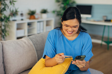 A young woman in a blue shirt is happily shopping online using her phone and a credit card, showcasing the convenience and joy of modern online shopping at home.