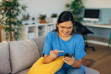 A smiling woman is using her smartphone and a credit card to make a purchase online from the comfort of her living room, blending shopping and technology effortlessly.