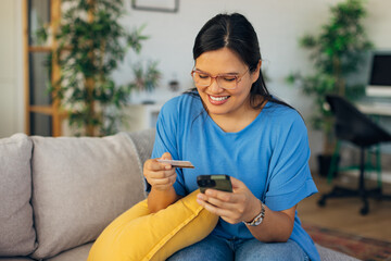 The image captures a smiling young woman comfortably using her smartphone and credit card to shop online from her stylish living room, highlighting modern consumerism.