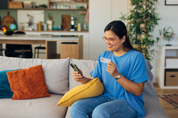 In a stylish home setting, a woman with glasses smiles while purchasing items online via smartphone, highlighting the blend of technology and comfort in daily living.