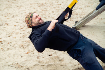Determined man working out with TRX suspension straps outdoors on sandy ground, showcasing strength and active lifestyle.