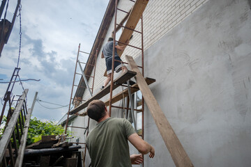 Two men work together on house renovation, with one on scaffolding installing a wooden board and the other assisting from below. This image captures home improvement and teamwork in construction.