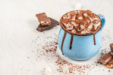 Hot chocolate with marshmallows in a blue mug topped with cocoa powder and chocolate drips, surrounded by chocolate pieces on a light background copy space for winter, cozy and festive food concepts