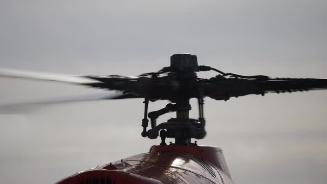 Close-up view of a red helicopter main rotor assembly in motion, showing spinning blades, rotor hub against an overcast sky, highlighting aerodynamics and rotational blur.