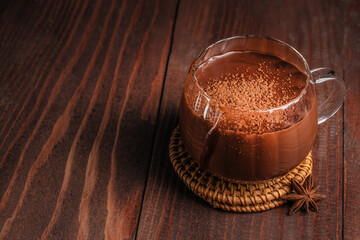 Rich hot chocolate in a clear glass mug topped with cocoa powder on a rustic wooden background copy space, creating a warm cozy dessert atmosphere perfect for winter drinks and sweet menu concepts