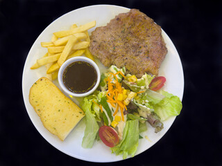 Thinly sliced ​​pork steak served with salad, garlic bread and french fries on a white plate.