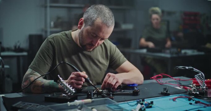 Worker With Tattoo on Arm Soldering Internal Electronics Onto Contemporary Drone Frame in Modern, Low Light Industrial Lab. Concept of Intersection of Industrial Design High Tech Assembly. Dolly Shot.