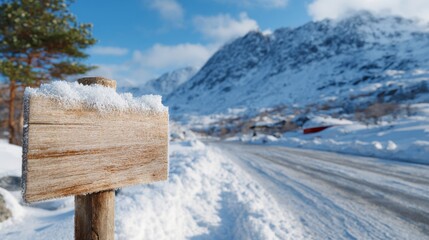 Fototapeta premium A rustic wooden signpost covered in snow stands beside a winding road, with majestic snow-capped mountains in the background.