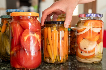 Hand reaching for pickled jars with assorted vegetables on kitchen counter. National Pickle Day