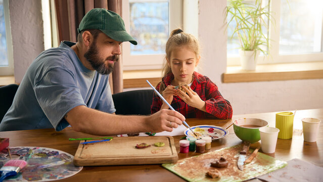 Father guiding daughter while painting clay figures at bright craft table. Concept of educational bonding, creative mentoring, practical learning routines, and collaborative hands-on activities.