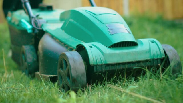 Gardener mowing grass with electric lawn mower wearing rubber boots. Man trimming green lawn using corded grass cutter in wellingtons. Worker cutting overgrown yard using powered mower while dressed