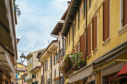A charming narrow street lined with colorful historic buildings, shuttered windows and flower-filled balconies on a bright day in Italy - Powered by Adobe