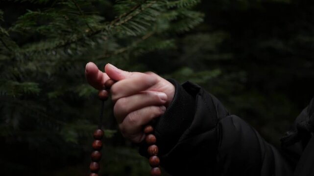 A close-up of a hand holding and running wooden rosary beads (prayer beads) against a blurred, dark green pine or fir forest background. The scene depicts spirituality, contemplation, and faith.