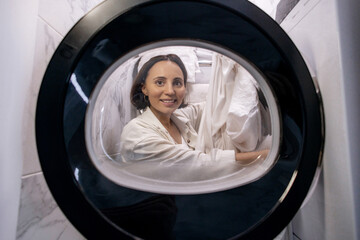 Fototapeta premium Smiling Woman Doing Laundry Seen from Inside Washing Machine Drum in Compact Bathroom with Marble Walls
