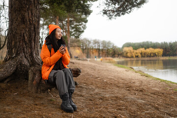 young Asian woman hiker sits resting near lake among the autumn forest.