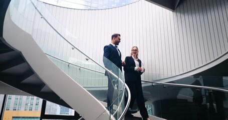 Two professional corporate colleagues walk down a spiral staircase in a modern office atrium while talking and holding coffee during a relaxed business break - Powered by Adobe