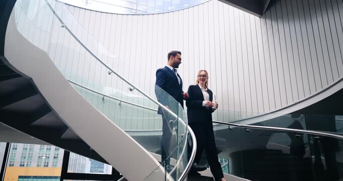 Two professional corporate colleagues walk down a spiral staircase in a modern office atrium while talking and holding coffee during a relaxed business break