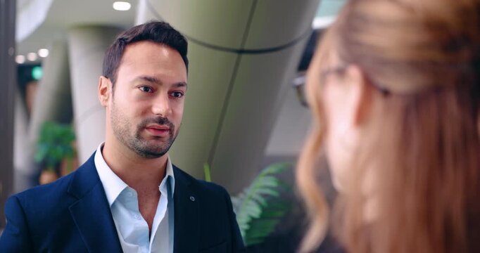 Face close up of professional businessman in suit talking seriously with female colleague seen over the shoulder in a modern corporate office hall during meeting