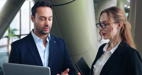 Confident CEO shows laptop screen to female employee while giving feedback on her work and discussing necessary improvements in a modern corporate office hallway