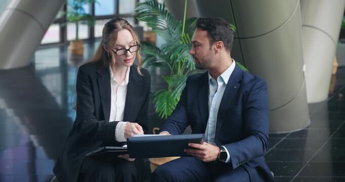 Experienced businessman sits with female employee giving constructive feedback while she takes notes on digital tablet in modern corporate office lobby during meeting