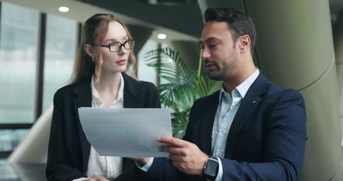 Professional male CEO gives detailed feedback to female employee while reviewing project paper documents and charts together in a modern corporate office