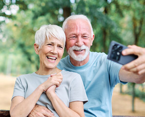Happy active senior couple having fun using smartphone, taking selfie, or having a video call and wearing sportswear, after having an exercise sport activity outdoors
