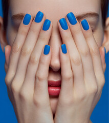 a close-up of the hands and nails, which cover her eyes with both palms while she smiles at the camera, showing blue nail polish on each finger. the background is white.