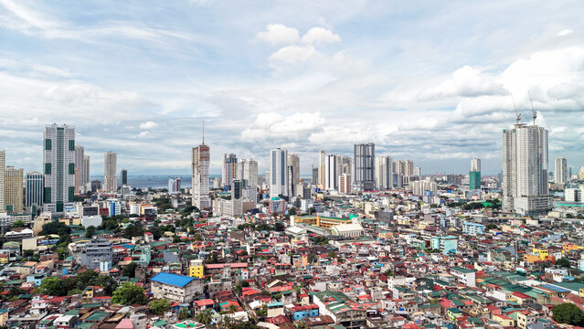 High angle drone view of Malate in Manila, Philippines, showing a dense patchwork of colorful low-rise houses in the foreground. Behind them rise tall modern condominium towers near Manila Bay, under 