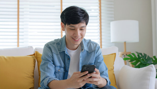 Young man smiling while using smartphone on couch at home, denim shirt, white tee, cozy living room, natural light, relaxed mood