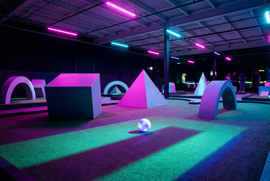 Indoor mini-golf course with vibrant neon lights. A futuristic playground with geometric obstacles illuminated in purple, pink, and green colors.