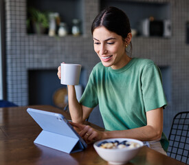 Portrait of a young woman using tablet or laptop computer, having online meeting or browsing internet, drinking coffee eating breakfast, drinking juice  in the kitchen at home