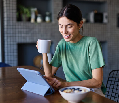 Portrait of a young woman using tablet or laptop computer, having online meeting or browsing internet, drinking coffee eating breakfast, drinking juice in the kitchen at home - Powered by Adobe