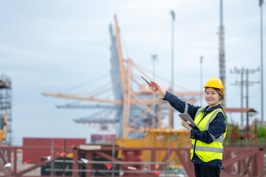 An Asian female engineer owns a company and monitors construction crane activities throughout the industrial port to maintain safe and accurate transportation processes, striving for development.