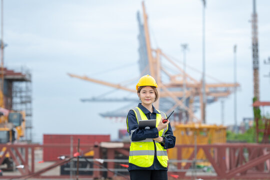 An Asian female engineer owns a company and evaluates transportation routes at an industrial port, coordinating construction crane schedules with laptop and radio technology.